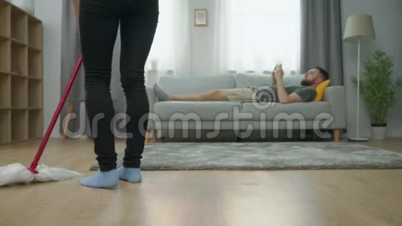 疲惫的妻子拖地板丈夫在沙发上玩智能手机视频的预览图