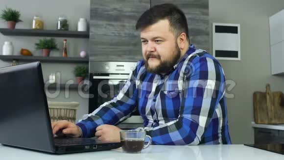 一个胖子坐在笔记本电脑前的厨房里高兴地喝着咖啡前面的厨房里有一只粗壮的雄性视频的预览图