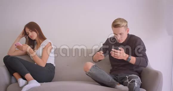 一对年轻可爱的白种人夫妇用手机坐在公寓里的沙发上的特写肖像男性视频的预览图