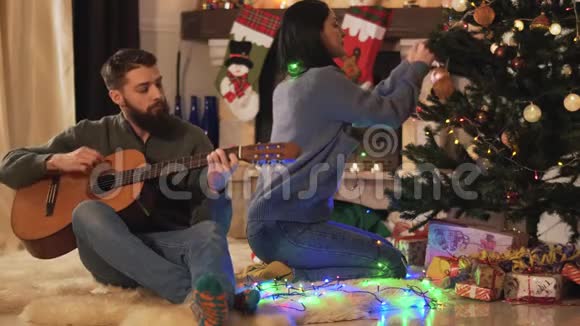 男人和女人坐在现代房间的圣诞树附近女孩挂玩具男人弹吉他快乐夫妇准备视频的预览图