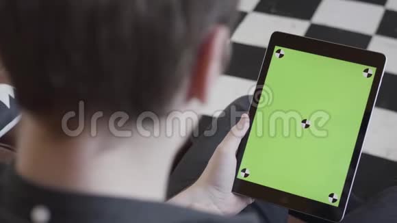 穿着深色牛仔裤的男人坐在沙发上手里拿着平板电脑黑白格子地板上有绿色屏幕视频的预览图