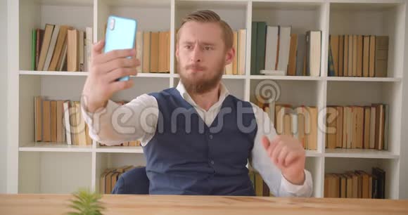 为年轻的白种人做生意的特写肖像在室内的工作场所带着书架在电话中自拍视频的预览图