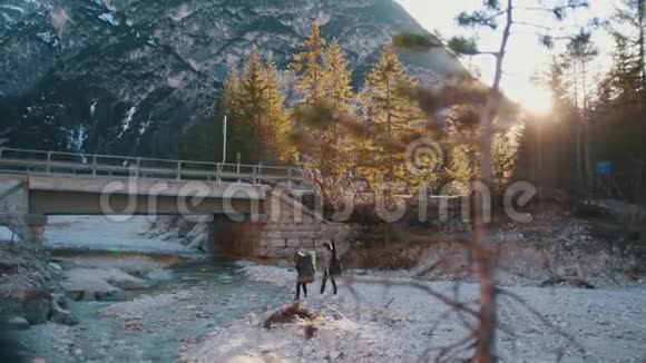 两个年轻女子背着背包和吉他在山上的一座桥下穿过岩石河视频的预览图