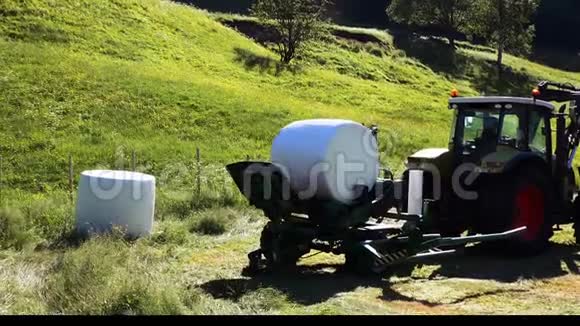 农夫用一台附在拖拉机上的机器把一个白色包裹的圆干草放在田边视频的预览图