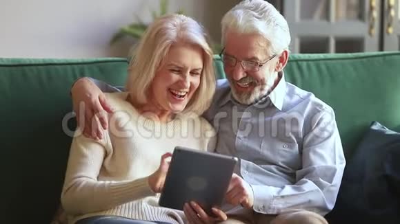 快乐的老两口用数码平板电脑在沙发上开怀大笑视频的预览图