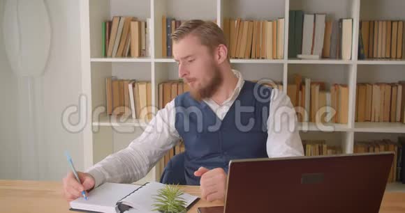 一位年轻的高加索商人用笔记本电脑拍摄了一幅特写肖像并在镜头中微笑着记笔记视频的预览图
