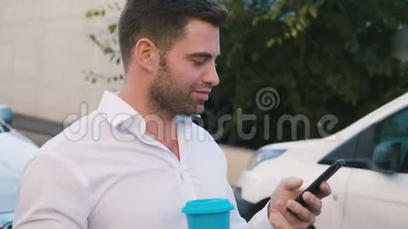 漂亮的商人站在办公楼附近的停车场上在他的智能手机上输入信息美味的饮料视频的预览图