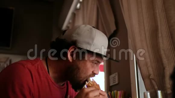 一个长着胡子戴着帽子的蓝眼睛嬉皮士在街头咖啡馆吃三明治汉堡在街头餐馆吃三明治汉堡视频的预览图