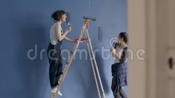 快乐的女性朋友一边唱歌一边用油漆滚筒在家里粉刷墙壁视频的预览图