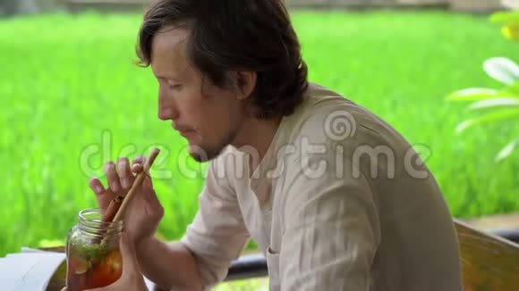 一个年轻人在一家有稻田背景的街边咖啡馆喝着薄荷和柠檬的冰茶的特写镜头视频的预览图