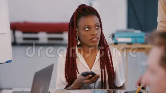 年轻漂亮的休闲非裔美国公司员工分心在团队会议办公室桌上使用智能手机视频的预览图