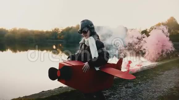 快乐的小飞行员女孩在日落湖上奔跑穿着有趣的飞机服装带着彩色烟雾镜头耀斑慢镜头视频的预览图