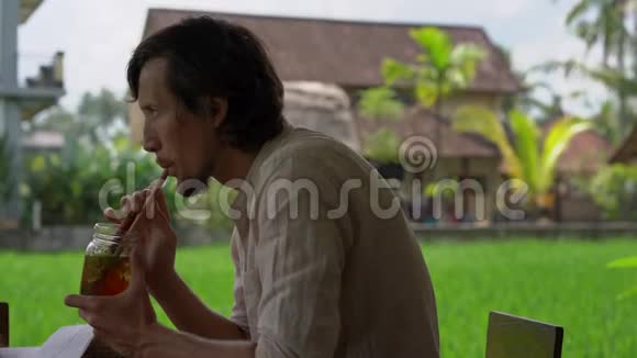 一个年轻人在一家有稻田背景的街边咖啡馆喝着薄荷和柠檬的冰茶的特写镜头视频的预览图