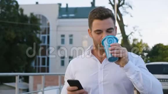 在办公大楼附近的停车场散步时帅哥使用智能手机和喝美味的咖啡看上去视频的预览图
