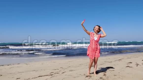 年轻可爱的女人戴着太阳镜站在沙滩上拍一张自拍照片背上有很强的波浪视频的预览图