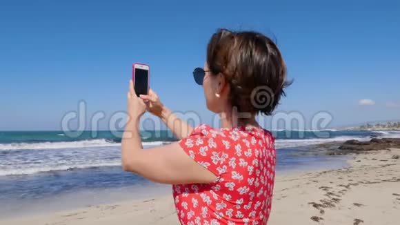 一个女人用手机站在海滩上拍照的特写镜头强风海浪背景上视频的预览图