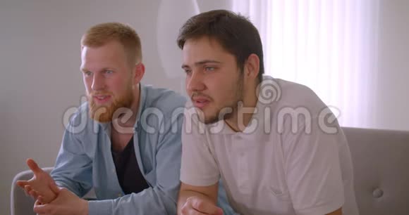 两位英俊的白种人成年男子在电视上兴奋地坐在沙发上观看体育节目的特写照片视频的预览图
