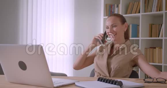 开朗的白种人女商人坐在笔记本电脑前在办公室里轻松地谈论手机视频的预览图