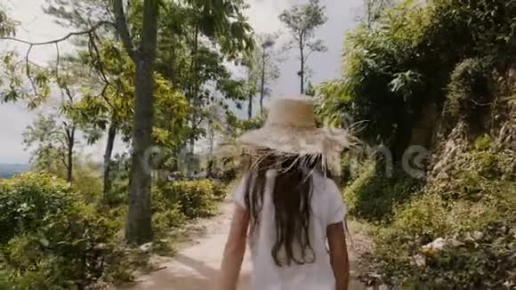 镜头跟随快乐的小57岁的女孩戴着大草帽在度假时沿着阳光明媚的热带森林道路散步视频的预览图