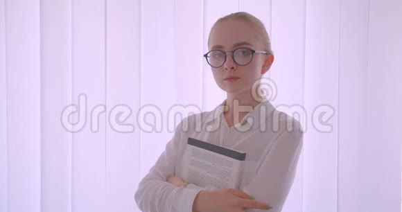 年轻漂亮的白种人金发女学生戴着眼镜拿着一本书看着摄像机站着的特写照片视频的预览图