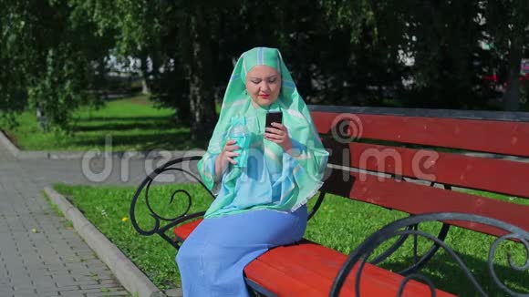 一位戴着浅色围巾的年轻穆斯林妇女在公园的长凳上下车喝着咖啡在信使里交谈视频的预览图