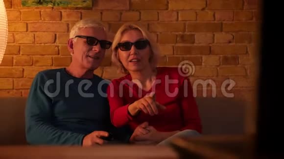 在舒适的公寓里坐在沙发上在电视上欢乐地观看3D电影的高级幸福夫妇的特写镜头视频的预览图