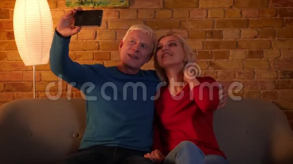 高年级幸福夫妇在电话里自拍的特写镜头欢快地坐在室内的沙发上视频的预览图