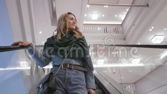 美丽的白种人女孩穿着蓝色夹克和绿色围巾微笑着在购物中心的自动扶梯下走去视频的预览图