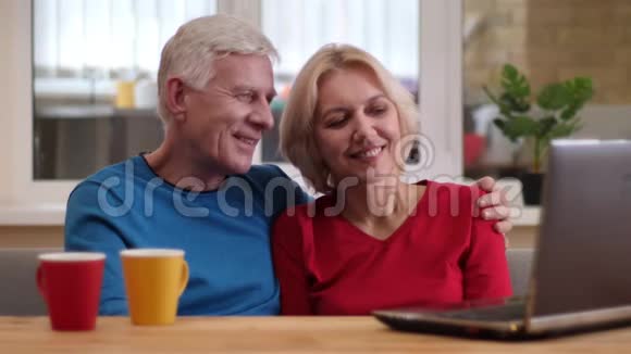 高级幸福夫妇的特写镜头他们用笔记本电脑在桌子上杯子里放着茶在舒适的室内愉快地微笑着视频的预览图
