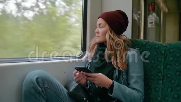 年轻孤独的白人沮丧的女孩戴着红帽子乘坐地铁或电车使用智能手机看着窗外视频的预览图