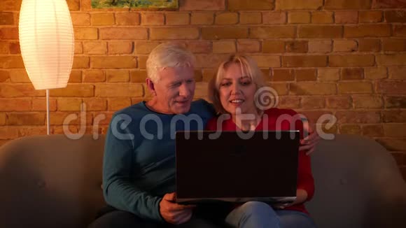 在笔记本电脑上使用社交媒体快乐地坐在室内沙发上的高级幸福夫妇的特写镜头视频的预览图