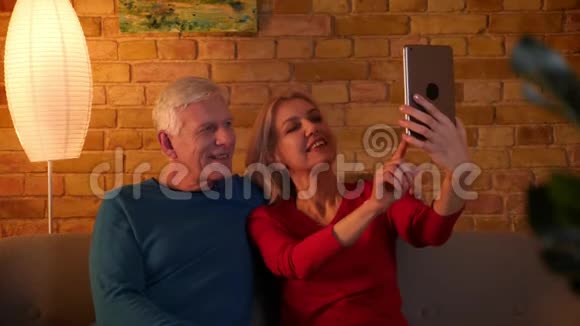 在舒适的公寓里坐在沙发上的高级幸福夫妇在平板电脑上自拍的特写镜头视频的预览图