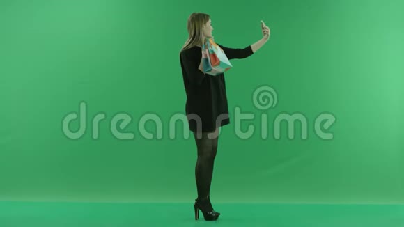 迷人的年轻女性在智能手机摄像头彩色钥匙绿色背景上用礼品袋拍照视频的预览图