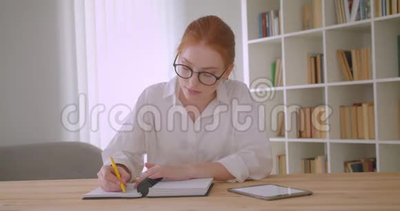 一位年轻漂亮的红头发女学生戴着眼镜用平板电脑写着一本笔记本坐在视频的预览图