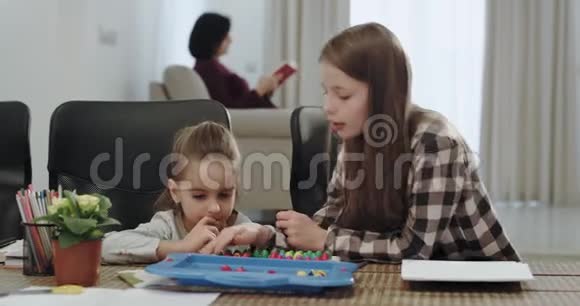 漂亮的两个女孩妹妹一起在厨房的桌子上玩耍而他们的祖母则在沙发上看书视频的预览图