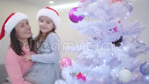 妈妈抱着女儿站在圣诞树附近可爱的女孩指着杉树枝上的一个粉红色的小树枝视频的预览图