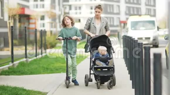 可爱的妈妈和她的儿子散步一个少年骑着滑板车妈妈带着婴儿车和一个蹒跚学步的男孩走着视频的预览图