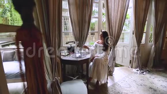 一个穿着白色连衣裙的黑发女人坐在桌子旁喝水背景中房间的老式内部视频的预览图
