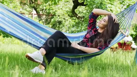 美丽快乐的年轻女子躺在花园的吊床上微笑着放松视频的预览图