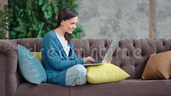 全镜头微笑的休闲年轻女性使用笔记本电脑打字坐在舒适的沙发上视频的预览图