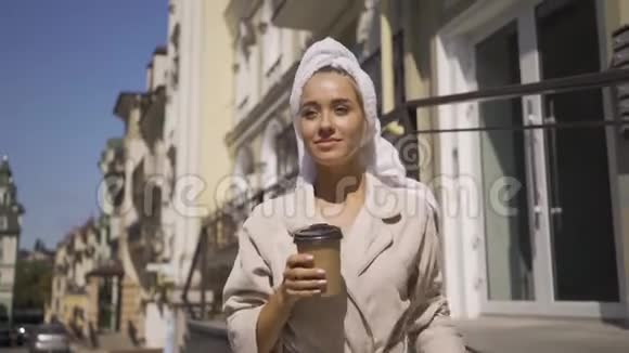 肖像微笑的年轻女子穿着浴袍头上戴着毛巾走在街上喝着咖啡自信的女孩视频的预览图