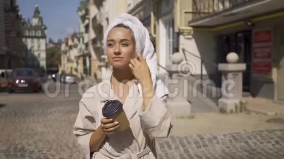 一幅美丽微笑的年轻女子穿着浴袍头上戴着毛巾走在街上喝着咖啡自信视频的预览图