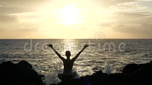 日落时分一个女人坐在岩石上观察海浪并在空中举起手臂的剪影电影慢动作视频的预览图