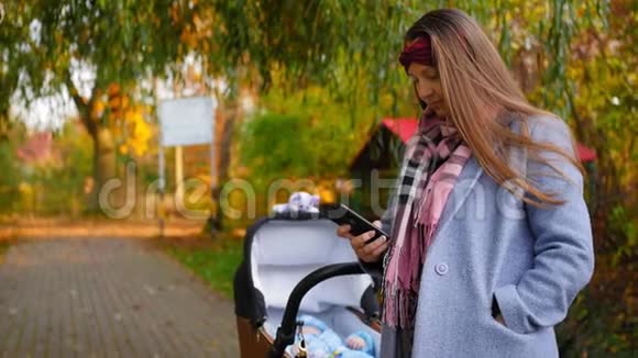 年轻的母亲手里拿着电话在公园秋天的婴儿车里和婴儿一起散步现代儿童视频的预览图