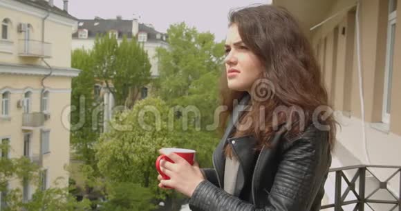 年轻的黑发女孩穿着皮夹克站在阳台上欣赏壮观的绿色城市景色喝着热茶视频的预览图