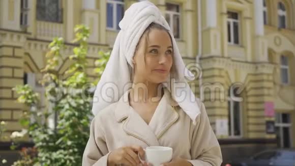 肖像微笑的年轻女子穿着浴袍头上有毛巾在街上喝咖啡自信的女孩享受着视频的预览图