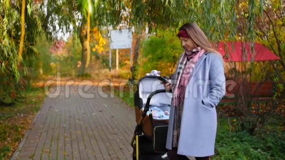 年轻的母亲手里拿着电话在公园秋天的婴儿车里和婴儿一起散步现代儿童视频的预览图
