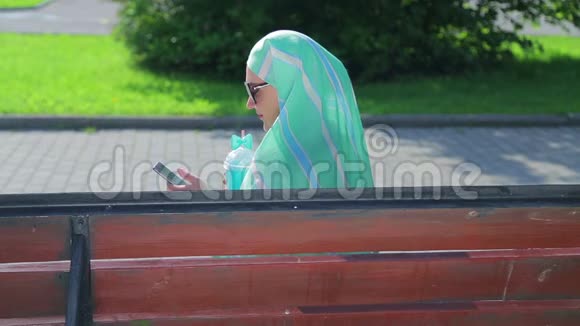 一位戴着浅色围巾的年轻穆斯林妇女坐在公园的长凳上喝着咖啡在信使里说话侧面视频的预览图