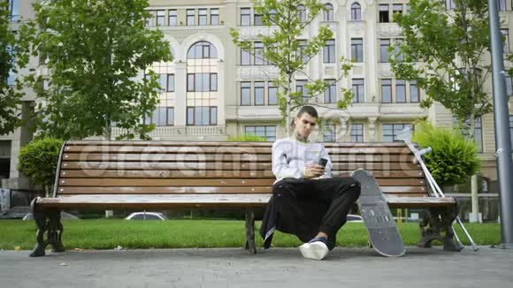 有魅力的男人坐在公园的长凳上用手机听音乐拐杖和滑板就在附近视频的预览图