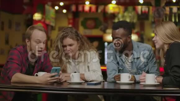 欢快的年轻朋友坐在咖啡馆里看智能手机的肖像各种种族的人坐在餐桌前视频的预览图